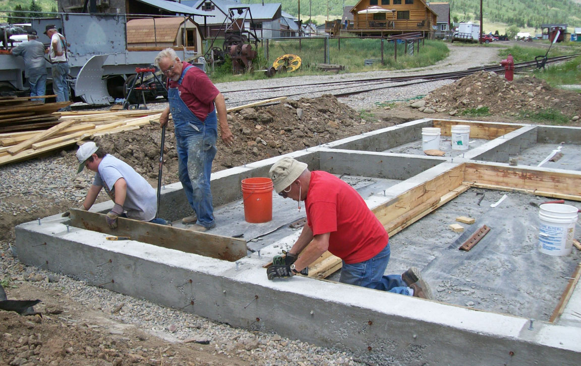 July 14. Joe Weigman, Jerry Hoffer, Ron Schlueter, and Judy Schlueter (taking the picture) removed forms from the oil shed foundation. In the background George Niederauer and Duane Danielson are working on the flanger.